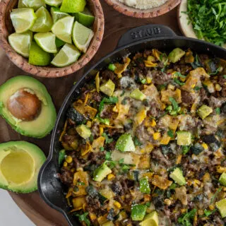 beef enchilada skillet with bowls of limes, cotija cheese, cilantro, and a halved avocado