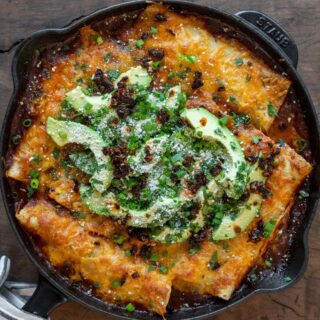 Tex Mex enchiladas in a cast iron skillet topped with sliced avocado, cilantro, green onion, salsa macha, and cotija cheese