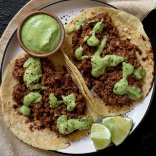 two ground bison tacos on a plate with lime wedges and a small wooden bowl filled with creamy jalapeño sauce